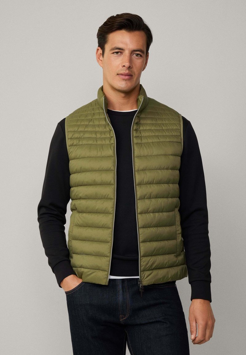 Gilet imbottito verde oliva, con zip, texture a righe orizzontali, colletto alto e tasche con zip nascoste, indossato sopra una maglietta nera.