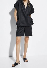 Svart kortärmad topp med dragkedja i halsringningen, kombinerad med matchande shorts. Båda plaggen har ett lätt, texturerat tyg. Svarta sandaler avslutar looken.