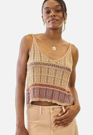 Mujer con top de punto beige y malva con patrones intrincados, combinado con pantalones beige de cintura alta sobre un fondo liso.