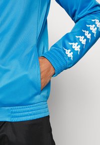 Modrá športová bunda z hladkého materiálu, so elastickými manžetami a vreckom. Biele logo na rukáve pridáva detail. Vidieť čierne šortky.