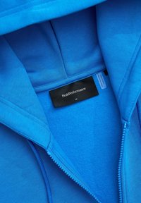 Jasnoniebieska kurtka z polaru z zamkiem błyskawicznym, na której widać metkę marki Peak Performance oraz metkę z rozmiarem medium wewnątrz kołnierza.