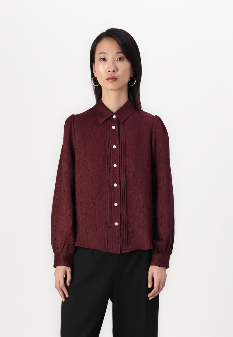 Burgundy long-sleeve blouse with a collared neck, button front, and pleated design. Features pearl-like buttons and soft texture.