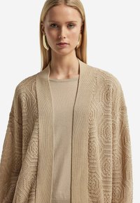 Beige öppen stickad cardigan med texturerade geometriska mönster och en solid stickad krage ovanpå. Bärs över en matchande beige topp.
