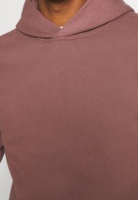 Bluza z kapturem w kolorze różanym, wykonana z miękkiego, teksturowanego materiału. Posiada zaokrąglony dekolt i minimalistyczne przeszycia. Gładki, prosty wzór bez zdobień.