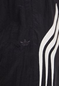 Nærbilde av svart Adidas-stoff med mørk brodert trefoil-logo og hvite signatur tre striper på siden.