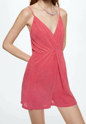 Femme portant une robe rose sans manches à décolleté en V avec un motif subtil et un collier translucide à perles, debout avec une main derrière le dos.