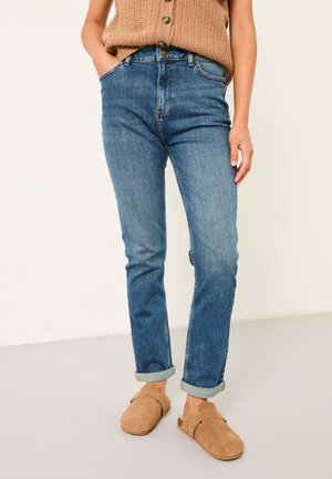 Person trägt hochgekrempelte blaue Jeans, beige Strickweste mit Knöpfen und beige Wildleder-Slipper und steht vor einer schlichten Wand.