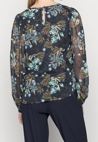 Blusa floreale in tessuto trasparente, con uno sfondo scuro e fiori verdi e blu, maniche lunghe a sbuffo e dettaglio a chiave sul retro.