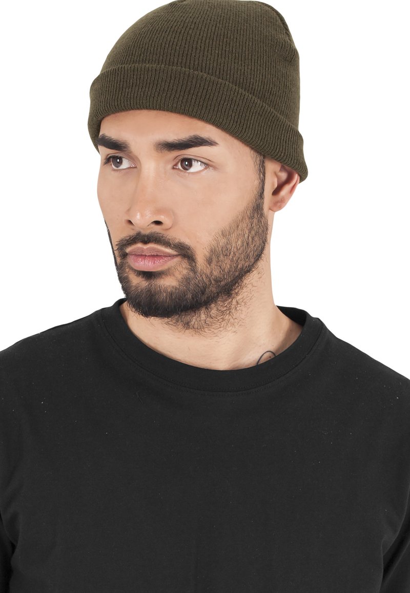 Grønn ribbestrikket lue iført med en svart crew neck t-skjorte. Nærbilde av ansiktet, som viser skjeggvekst og en nøytral uttrykk.