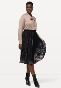 Beige blouse met strikdetail en lange mouwen, gecombineerd met een geplooide zwarte rok met een multicolore binnenvoering, aangevuld met zwarte enkellaarzen.