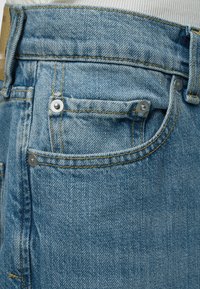 Κοντινή όψη τσεπης μπλε τζιν με μεταλλικά καρφιά και κίτρινη ραφή σε λευκό υφασμάτινο φόντο.