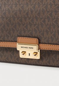 Ruskeakuvioinen synteettinen käsilaukku, jossa on kultainen metallilukko ja pienennetty logokuva. Muotoilussa on sileä nahkainen yksityiskohta.