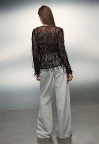 Black sheer top with hanging fringe and beaded accents, paired with light gray wide-leg trousers. Textured fabric with a layered look.