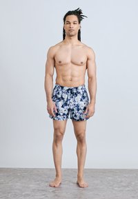 Άνδρας με μακριές dreadlocks που φοράει μπλε και λευκό μαγιό tie-dye, που στέκεται ξυπόλυτος σε γκρι δάπεδο μπροστά σε ομοιόμορφο ανοιχτό φόντο.