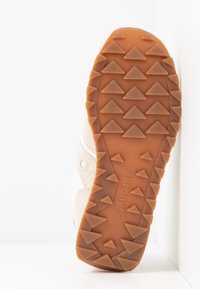 Baskets de couleur claire avec une surface texturée, possédant une semelle en caoutchouc avec des crampons triangulaires et une marque. Design épuré, pratique pour l'adhérence.
