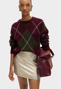 Suéter de argyle burdeos y verde con mangas enrolladas, combinado con una falda corta metálica dorada y un bolso de cuero burdeos. Texturas suaves.