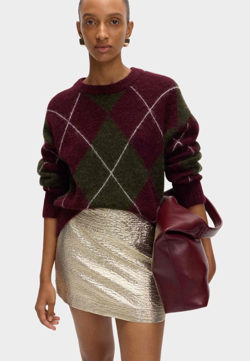 Suéter de argyle burdeos y verde con mangas enrolladas, combinado con una falda corta metálica dorada y un bolso de cuero burdeos. Texturas suaves.