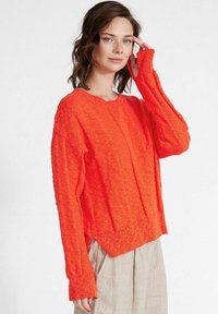 Maglione lavorato a maglia arancione brillante con una finitura testurizzata, scollo tondo e maniche lunghe leggermente sovradimensionate. Presenta un orlo corto.