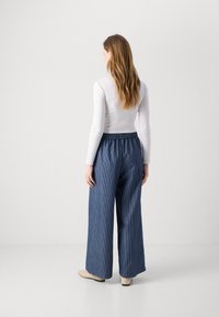 Široke denim hlače z elastičnim pasom, ki imajo vzorec s punčico. Model nosi oprijet belo majico z dolgimi rokavi in bež čevlje.