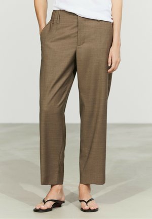 Persona che indossa pantaloni marroni a gamba larga abbinati a sandali neri con tacco, in piedi su un pavimento liscio grigio chiaro.