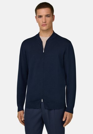 Junger Mann, der eine marineblaue Strickjacke mit Reißverschluss über einem beigen Hemd und navyblauen Hosen trägt, steht vor einem schlichten hellen Hintergrund.