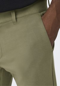 Pantalon vert olive ajusté avec une texture lisse, comprenant des poches latérales et une taille nette, partiellement montré avec la main d'un mannequin.