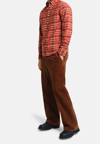 Camicia a quadri rossa con bottoni e dettagli bianchi, abbinata a pantaloni in velluto a coste marroni e scarpe slip-on nere. Vestibilità comoda con tasche anteriori.