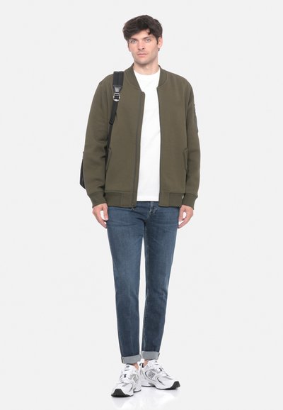 Giacca bomber oliva, t-shirt bianca, jeans blu e sneakers bianche. La giacca presenta una chiusura con zip e tasche laterali. Design minimalista.