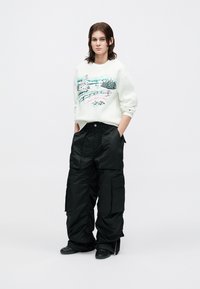 Jovem com cabelo até aos ombros a usar um sweatshirt branco com uma estampa de paisagem de inverno e calças cargo pretas largas, com as mãos nos bolsos.