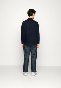 Camisa de manga larga azul marino con un pequeño cuello, puños abotonados y un pliegue en la parte posterior, combinada con pantalones de mezclilla oscuros de corte holgado y zapatillas blancas.