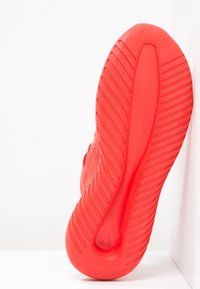 Červená gumová podrážka tenisky s diagonálními drážkami, zaoblenými okraji a logem blízko paty. Design se zaměřuje na trakci a odolnost.