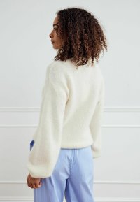 Kreminio atspalvio, pūkuotas megztinis su aukštu apykakle ir burbulinėmis rankovėmis, derinamas su mėlynomis dryžuotomis kelnių, matomas iš nugaros.