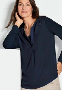 Marineblaues Langarmblusenshirt mit einem Schalkragen, gefältelten Schultern und einer fließenden Passform; kombiniert mit hellen Hosen.