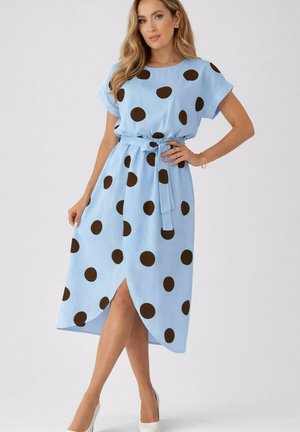 Femme posant dans une robe midi bleu clair à grands pois noirs, taille ceinturée, manches courtes et talons hauts blancs.