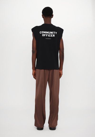 Camisa preta sem mangas com "OFICIAL DA COMUNIDADE" em letras grandes brancas nas costas, combinada com calças largas de tom castanho claro. Textura suave.