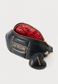 Juodas odinis fanny pakas su raudonu raštuotu interjeru, auksinėmis detalėmis ir širdies formos užtrauktuku, ant kurio užrašyta "LOVE MOSCHINO" auksinėmis raidėmis.