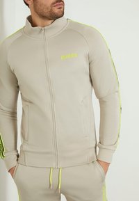 Giacca zip-up beige chiaro con accenti giallo fluorescente e logo. Presenta un colletto in piedi, polsini a coste e tasche laterali.