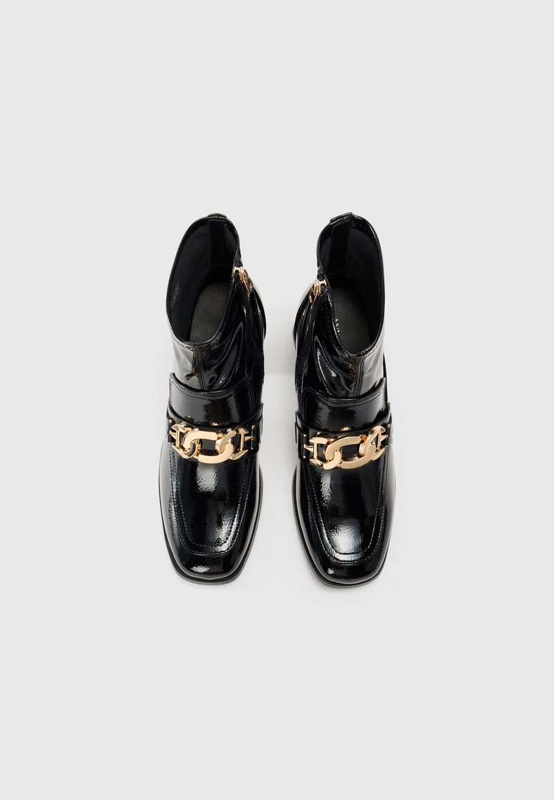 Botas de tobillo de charol negro con punta cuadrada, detalle de cadena dorada y cremalleras laterales. Textura suave con acabado brillante.
