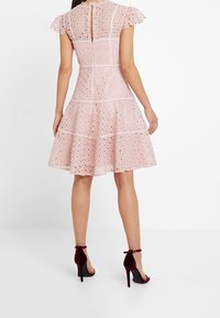 Vaaleanpunainen mekko, jossa on reikäkangasta, hihat, ja liehuva helma. Takana avaus, jossa on avainreikä ja kontrastiväriset valkoiset saumat. Yhdistetty marooninvärisiin korkokenkiin.