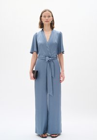 Blaue, weit geschnittene Jumpsuit mit Wickeldesign, kurzen Ärmeln und einem gebundenen Taillengürtel. Glattes Gewebe mit einer subtilen Textur; kombiniert mit einer schwarzen Clutch.