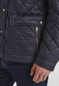 Gepolsterte, marineblaue Daunenjacke mit einer vorderen Taschenklappe, einem Reißverschluss-Seitentasche und gerippten Bündchen. Glatte, glänzende Stofftextur.