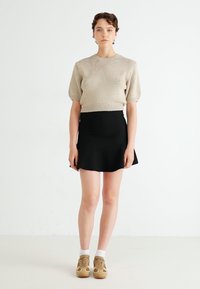 Beige gestrickter Pullover mit kurzen Ärmeln und strukturiertem Design, kombiniert mit einem schwarzen, ausgestellten Minirock. Das Modell trägt braune Sneaker mit weißen Socken.