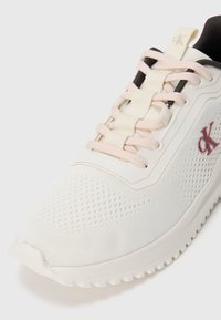 White and light pink sneaker with perforated fabric, thick sole, black inner lining, and red "cK" logo on the side.