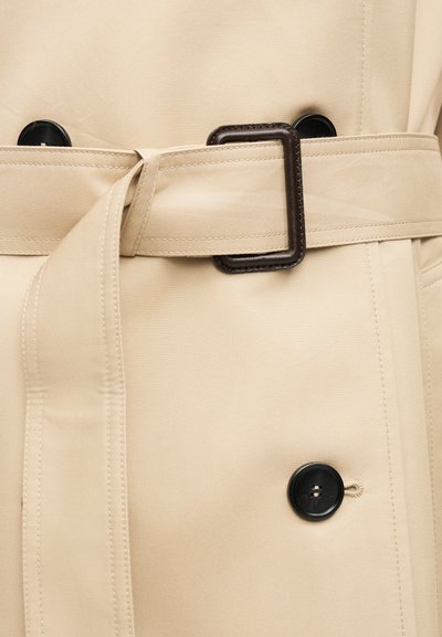 Μπεζ παλτό τύπου trench με ραμμένη ζώνη που περνά μέσα από μια σκούρα ορθογώνια αγκράφα, με μαύρα κουμπιά μπροστά.