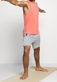 Koronkowy top w kolorze koralowym i szare szorty noszone przez osobę w pozie jogi na macie. Top jest bez rękawów z bocznymi rozcięciami; szorty sięgają powyżej kolan.