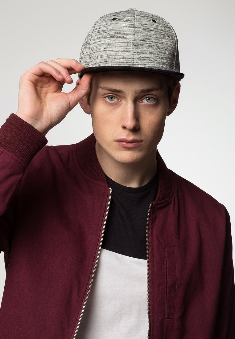 Sivá čiapka s textúrovým, šumeným vzorom. Model má na sebe marónovú bomber bundu cez čierno-biele tričko, ruku položenú na okraji čiapky.