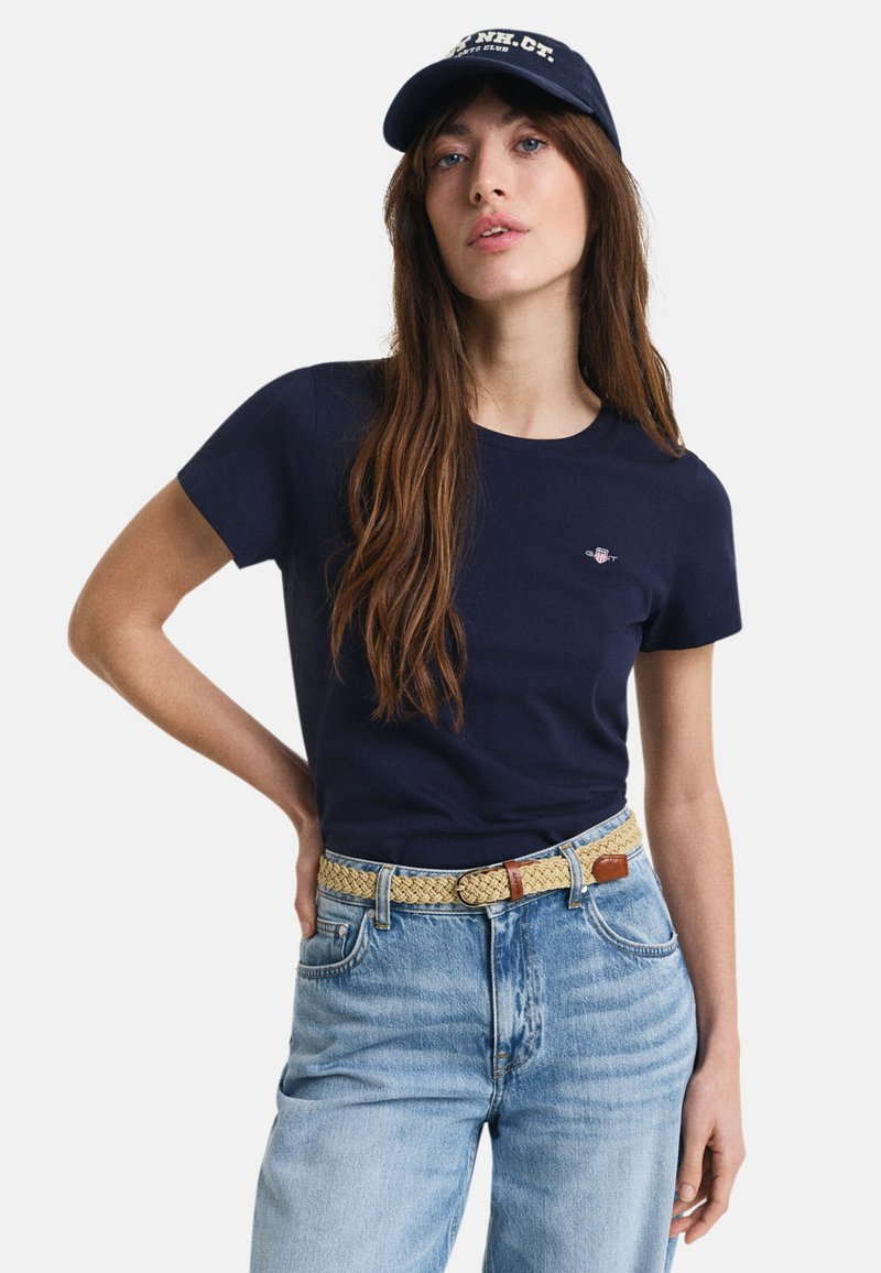 Marinblå bomullströja med korta ärmar, prydd med ett litet emblem på bröstet. Kombinationen med ljusblå jeans och ett vävt beige bälte.