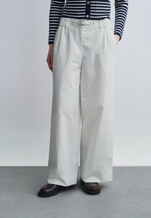 Žena v bielych nohaviciach s vysokým pásom a širokými nohavicami, s bielym opaskom, modro-bielej pruhovanej blúzke s dlhým rukávom a tmavých mokasínach.