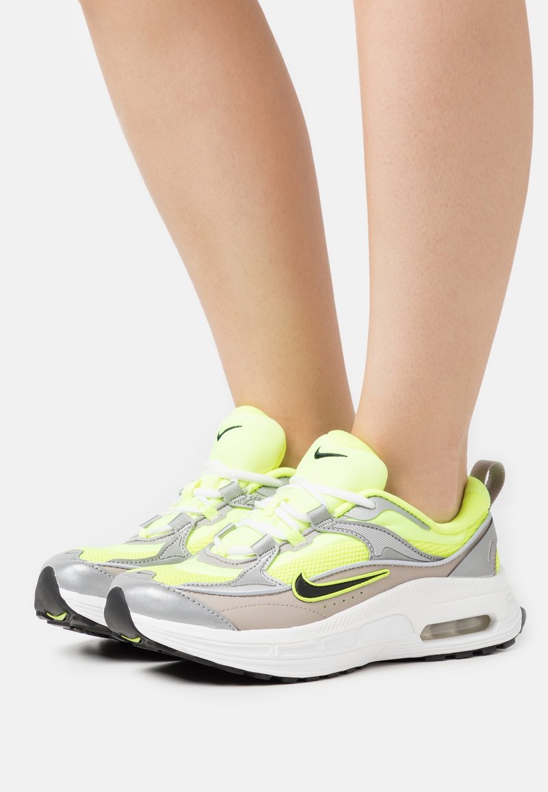 Jambes portant des baskets Nike Air Max jaune néon et gris avec des lacets blancs et des logos Swoosh noirs sur fond blanc.