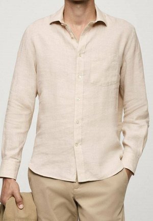 Man die een lichtbeige lange mouwen overhemd met knopen en een kaki broek draagt, met een beige hoed in één hand, staand tegen een effen achtergrond.